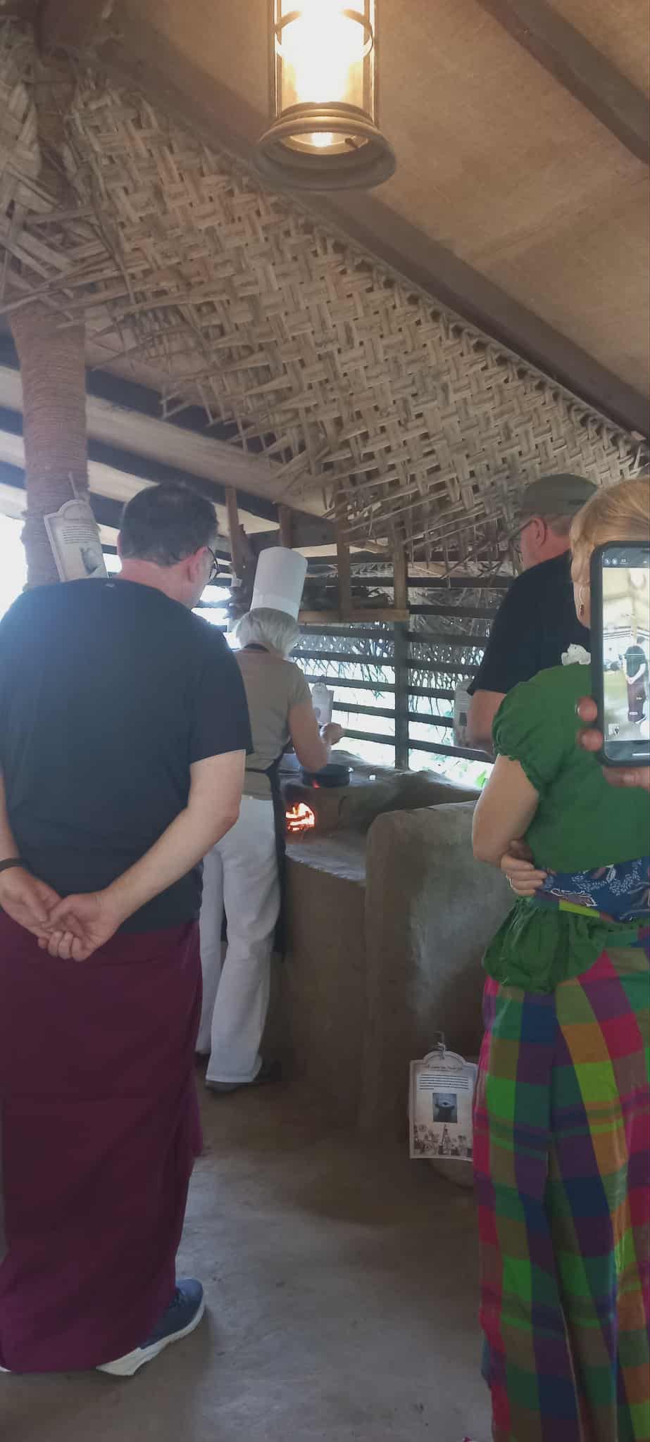 Guests watching a demonstration under a woven roof in a village