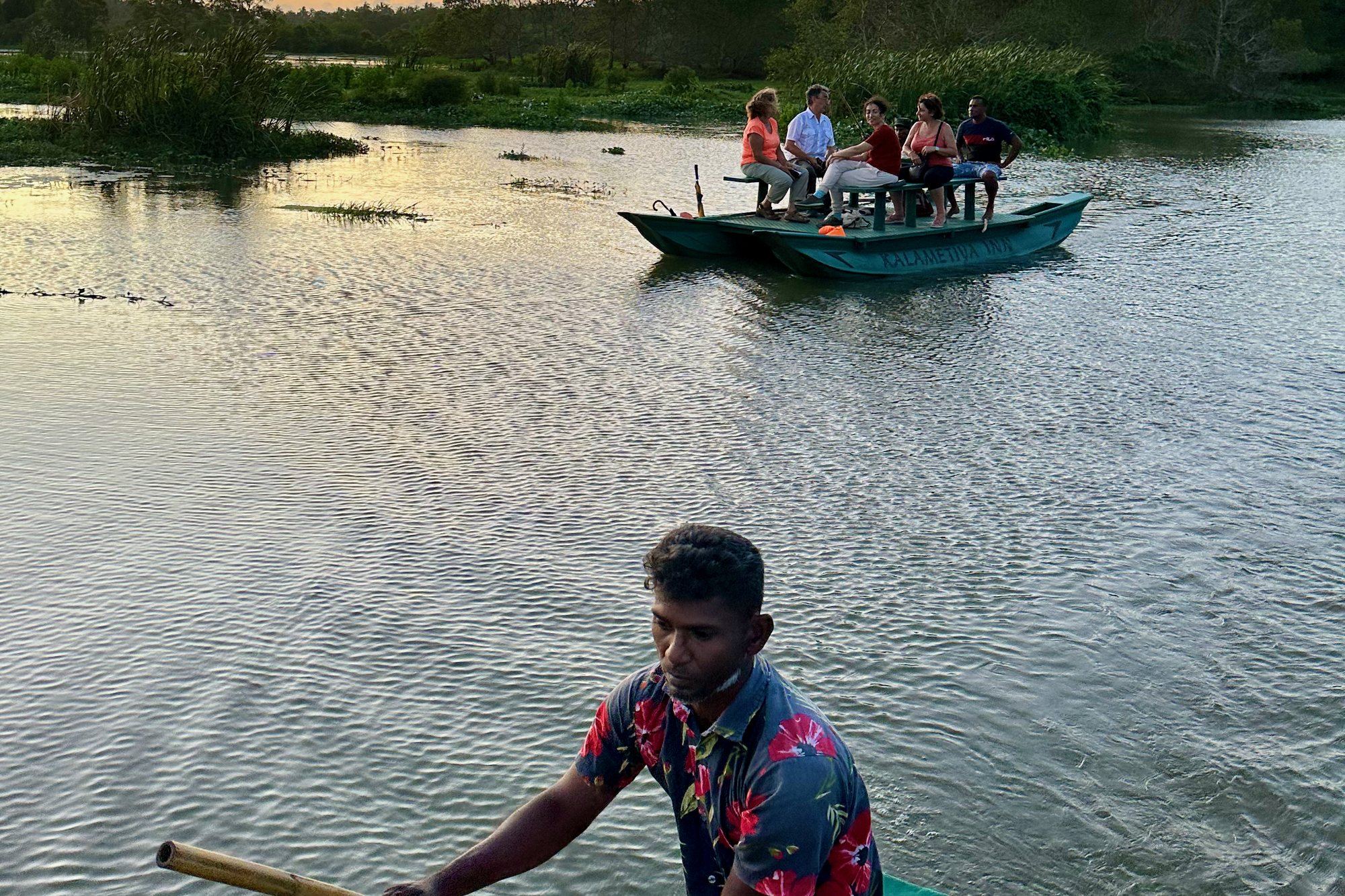 A boatman poling a small boat across the Kalametiya lagoon at sunset, with guests on a second boat behind
